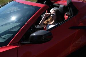 luxueux voiture et fille une fille est assis derrière le roue de une rouge voiture avec sa mains en haut, jouit vie, une riches vie, une lot de argent. photo