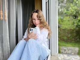 une fille dans une blanc chemise une adolescent est assis sur une blanc rebord de fenêtre le vue de le rue elle pouvez être vu dans le fenêtre dans sa mains elle détient une blanc iphone 13 pro max elle regards à le écran calmement photo