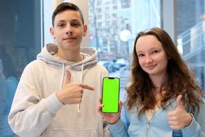 souriant gingembre longue cheveux adolescent garçon et fille montrant horizontal téléphone avec vert écran chrominance clé et parlant mots à maison, produit présentation, les adolescents application, Jeu et en ligne magasin publicité. photo