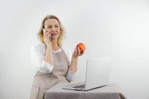 femme parlant sur téléphone est dérangé et mécontent dans mains elle a rouge Pomme diététique correct nutrition menu recettes dans de face de portable dans mains téléphone tablier sur blanc Contexte âge moyen femme blond photo