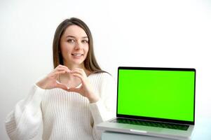 Jeune femme parler avec copains sur appel et spectacles cœur avec mains sur caméra de Accueil bureau. virtuel Noël fête réunion équipe télétravail. éloigné conférence ordinateur vert écran voir. photo