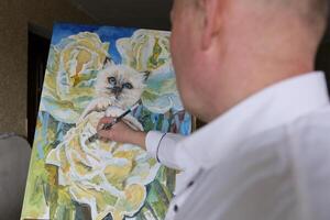 le artiste homme avec une brosse dessine à une blanc chat avec bleu yeux sourit regards dans le Cadre le maîtrise main La peinture dessin fleurs brillant délicieux beauté sur le mur décoration art photo