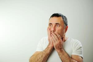 négatif émotion horreur surprise en gardant votre secret. homme couvrant bouche avec main le homme couvert le sien bouche avec le sien mains et ouvert le sien yeux large noir cheveux avec gris poilu bras blanc T-shirt photo