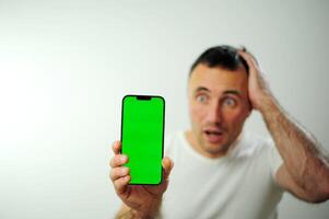 Nouveau les technologies gagner adulte téléphone vert écran chromakey surpris et en portant le sien main derrière le sien tête il est très inquiet une blanc T-shirt et une blanc Contexte pour une homme avec foncé cheveux photo