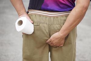 proche en haut homme détient tissu papier rouleau et main toucher le sien bas. concept, avoir besoin à aller à toilettes. diarrhée. les hémorroïdes. santé problèmes et santé se soucier. photo