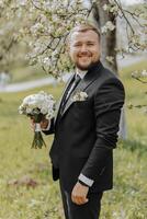 le marié, habillé dans une noir costume, des stands dans la nature avec une bouquet de fleurs dans le sien main. un élégant homme dans une classique noir costume. portrait de une homme photo