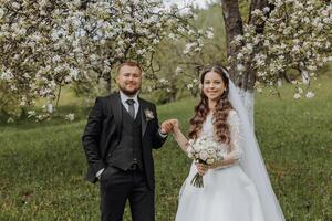 le jeune marié détient le main de le sien la mariée contre le Contexte de une épanouissement arbre dans une vert jardin dans printemps photo