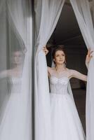 une femme dans une blanc robe est permanent dans de face de une fenêtre, à la recherche en dehors photo