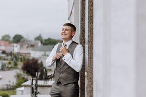 une homme dans une costume et gilet est permanent sur une balcon, ajustement le sien attacher photo