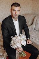 une homme dans une costume est séance sur une lit en portant une bouquet de fleurs photo