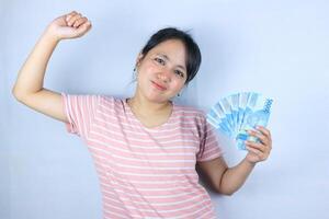 portrait de une souriant asiatique Jeune femme en portant en espèces Rupiah et célébrer sur blanc Contexte photo