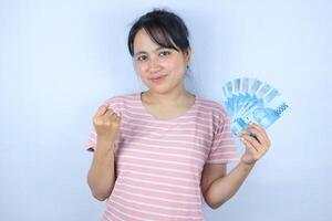 portrait de une souriant asiatique Jeune femme en portant en espèces Rupiah et célébrer sur blanc Contexte photo