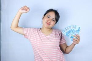 portrait de une souriant asiatique Jeune femme en portant en espèces Rupiah et célébrer sur blanc Contexte photo