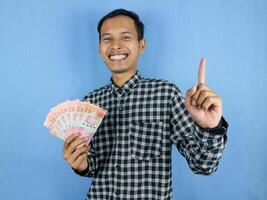 excité expression Beau asiatique homme tenir papier argent billets de banque avec montrer du doigt en haut geste photo