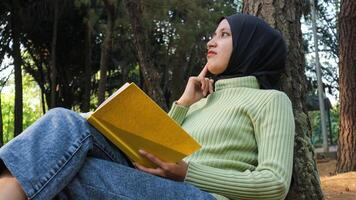 portrait de Jeune musulman femme portant hijab en train de lire une livre tandis que penché contre une arbre dans le parc photo