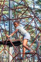 joyeux fille grimpe une corde structure à une parc, sa sourire exprimer le frisson de aventure au milieu de grand pins, parfait pour thèmes de actif enfance, et des sports activités. photo