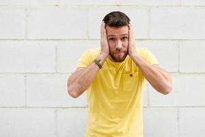 jeune homme drôle sur le mur de briques avec une drôle d'expression couvrant ses oreilles. photo