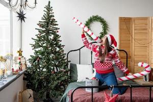 Jeune femme en pull rouge et bonnet de Noel dansant sur le lit pour célébrer Noël photo