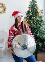 Jeune femme en bonnet de noel tenant une boule disco miroir debout près de l'arbre de Noël