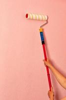 un rouleau à peinture laineux dans les mains féminines d'un peintre sur fond de mur rose.