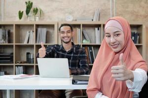 portrait d'un entrepreneur en démarrage d'entreprise, d'un jeune homme et d'une belle femme propriétaire, de deux partenaires islamiques, regardant la caméra, souriant joyeusement et levé le pouce dans un petit bureau de commerce électronique. photo