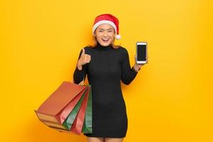 souriante jeune femme asiatique en chapeau de père noël tenant un téléphone portable à écran blanc et gesticulant les pouces vers le haut, tenant des sacs à provisions isolés sur fond jaune photo