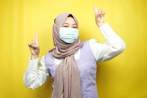 femme musulmane portant un masque médical, main pointant vers un espace vide, main pointant vers le haut présentant quelque chose, isolée sur fond jaune