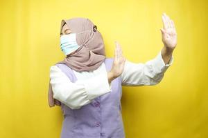 femme musulmane portant un masque médical avec la main rejetant quelque chose, la main arrêtant quelque chose, la main n'aimant pas quelque chose, isolée sur fond jaune photo
