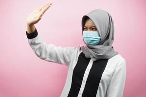 femme musulmane portant un masque médical avec la main rejetant quelque chose, la main arrêtant quelque chose, la main n'aimant pas quelque chose, isolée sur fond rose photo