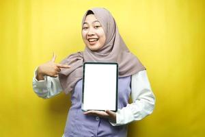 belle jeune femme musulmane asiatique souriante, excitée et joyeuse tenant une tablette avec un écran blanc ou vierge, faisant la promotion de l'application, faisant la promotion du produit, présentant quelque chose, isolée sur fond jaune photo