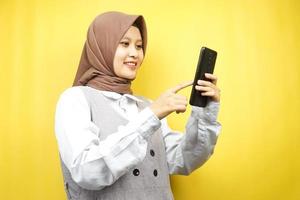 belle jeune femme musulmane asiatique souriant avec confiance tenant un smartphone isolé sur fond jaune, concept publicitaire photo