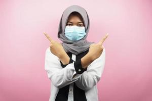 femme musulmane portant un masque médical, main pointant vers un espace vide, main pointant vers le haut présentant quelque chose, isolée sur fond rose