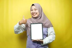 belle jeune femme musulmane asiatique souriante, excitée et joyeuse tenant une tablette avec un écran blanc ou vierge, faisant la promotion de l'application, faisant la promotion du produit, présentant quelque chose, isolée sur fond jaune photo