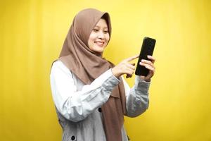 belle jeune femme musulmane asiatique souriant avec confiance tenant un smartphone isolé sur fond jaune, concept publicitaire photo