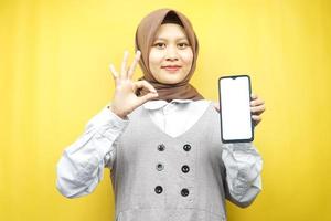 belle jeune femme musulmane asiatique souriante confiante, enthousiaste et joyeuse avec une main tenant un smartphone, faisant la promotion de quelque chose, faisant la promotion de l'application, isolée sur fond jaune photo