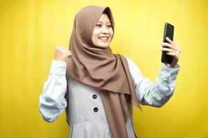 belle jeune femme musulmane asiatique souriante confiante, enthousiaste et joyeuse avec les mains tenant le smartphone, serrer les mains, poinçonner, victoire, succès, bon travail, isolé sur fond jaune