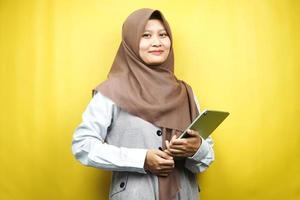 belle jeune femme musulmane asiatique souriante, excitée et joyeuse tenant une tablette, isolée sur fond jaune