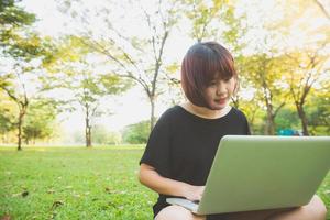 les jambes de la jeune femme asiatique sur l'herbe verte avec un ordinateur portable ouvert. les mains de la fille sur le clavier. concept d'apprentissage à distance. heureux hipster jeune femme asiatique travaillant sur ordinateur portable dans le parc. étudiant étudiant à l'extérieur.