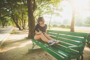 Happy hipster young asian woman écrit dans son journal dans le parc. heureux hipster jeune femme asiatique travaillant sur ordinateur portable dans le parc. étudiant étudiant à l'extérieur. concept de plein air femme mode de vie. lumière évasée. photo