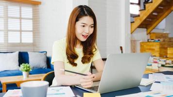 Une femme d'affaires asiatique utilisant un ordinateur portable parle à ses collègues du plan lors d'un appel vidéo tout en travaillant de manière intelligente à domicile dans le salon. auto-isolement, distanciation sociale, quarantaine pour la prévention du virus corona.