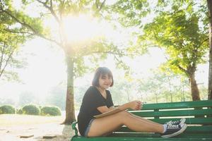 Happy hipster young asian woman écrit dans son journal dans le parc. heureux hipster jeune femme asiatique travaillant sur ordinateur portable dans le parc. étudiant étudiant à l'extérieur. concept de plein air femme mode de vie. lumière évasée. photo