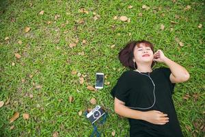 jeune femme asiatique allongée sur l'herbe verte écoutant de la musique dans le parc avec une émotion froide. jeune femme se reposant sur l'herbe avec sa liste de lecture musicale. activité de plein air dans le concept de parc. photo