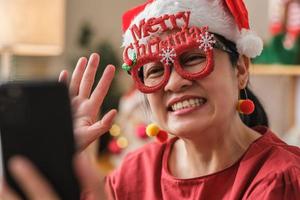 une femme asiatique porte une soirée d'appel vidéo avec un chapeau de père Noël avec des amis avec un téléphone portable sur une table à la maison photo