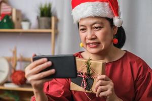 une femme asiatique porte une soirée d'appel vidéo avec un chapeau de père Noël avec des amis avec un téléphone portable sur une table à la maison photo