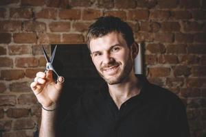 Le coiffeur masculin de portrait tient des ciseaux pointus et souriant contre un fond de mur de briques photo