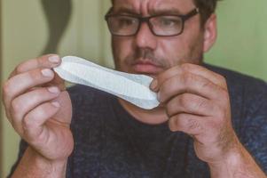 un homme avec des lunettes regarde une serviette hygiénique féminine avec surprise photo