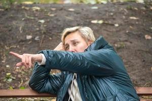 une fille dans une veste sombre est assise sur un banc dans le contexte de la nature automnale pointant vers le côté avec sa main pensant et regardant vers l'avenir photo