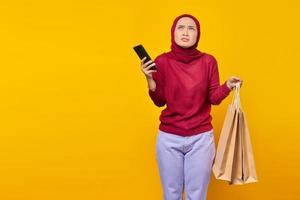 Jeune femme asiatique pensive tenant un téléphone portable et des sacs à provisions sur fond jaune photo