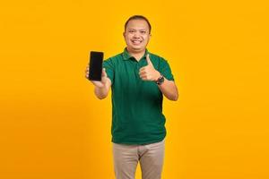jeune homme asiatique gai montrant un téléphone portable et montrant un geste de pouce levé sur fond jaune photo