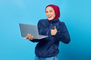belle femme asiatique utilisant un ordinateur portable souriant et montrant un signe de pouce levé sur fond bleu